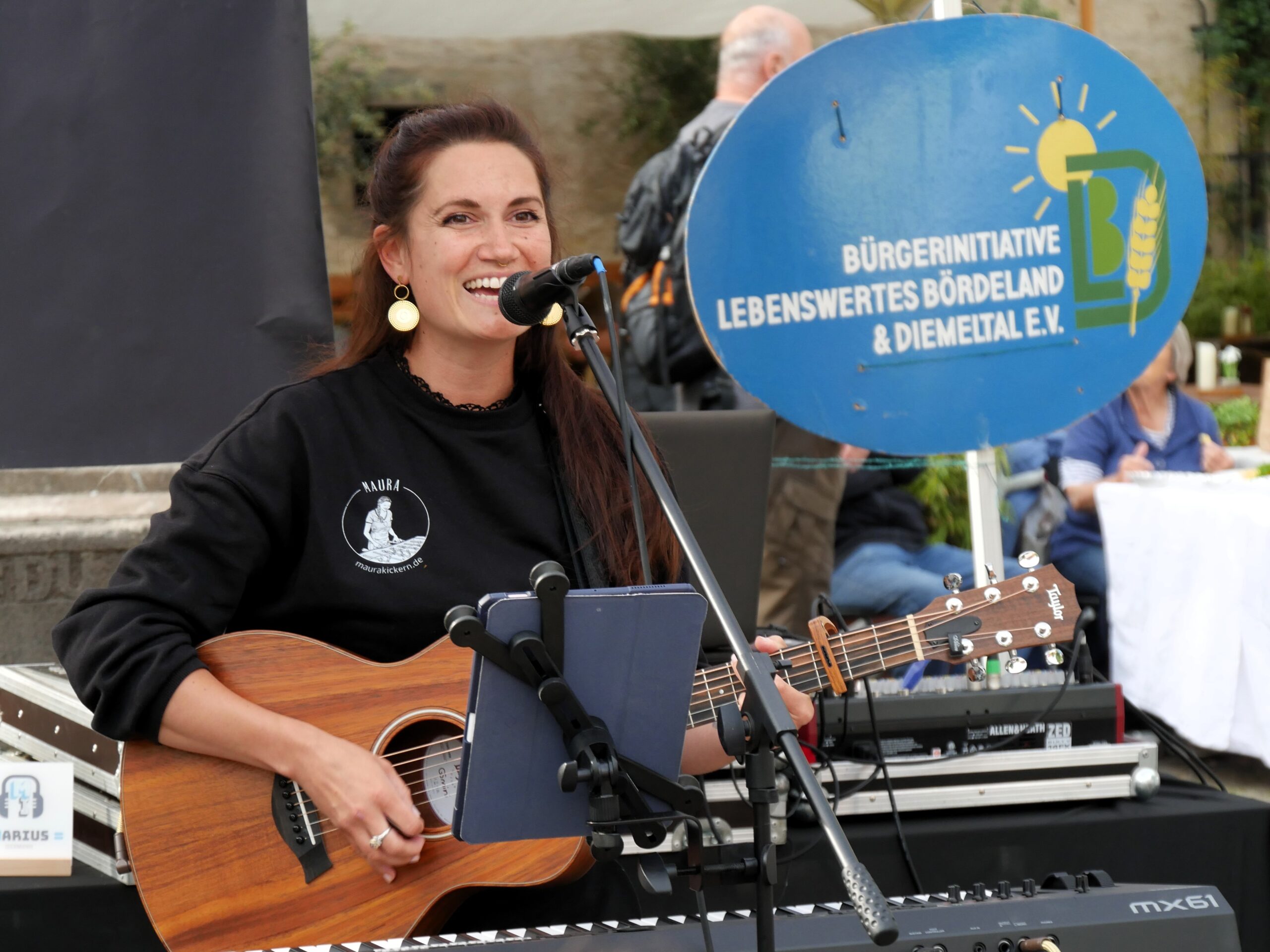 Sängerin MAURA mit Gitarre vor dem hellblauem Logo der Bürgerinitiative Lebenswertes Bördeland & Diemeltal.